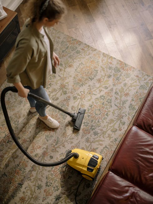 A woman with curly hair, wearing a khaki jacket, blue jeans, and white sneakers, is performing surface cleaning on a patterned area rug in a residential living room. She is holding a vacuum cleaner ho
