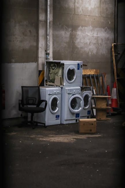 A large pile of old, disused computer monitors and electronic devices stacked haphazardly against a wooden wall, with some devices showing dusty or dirty surfaces. The monitors are of various shapes a