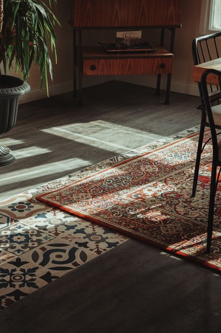 Close-up view of a traditional patterned area rug with intricate floral motifs in shades of red, blue, yellow, and cream, placed on a polished wooden floor with a natural light finish. The edge of a d