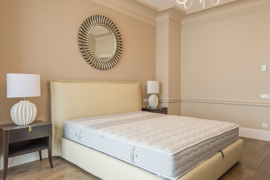A modern bedroom featuring a large double bed with a light grey upholstered headboard and a white mattress, positioned against a plain white wall. To the right of the bed, there are two hanging pendan